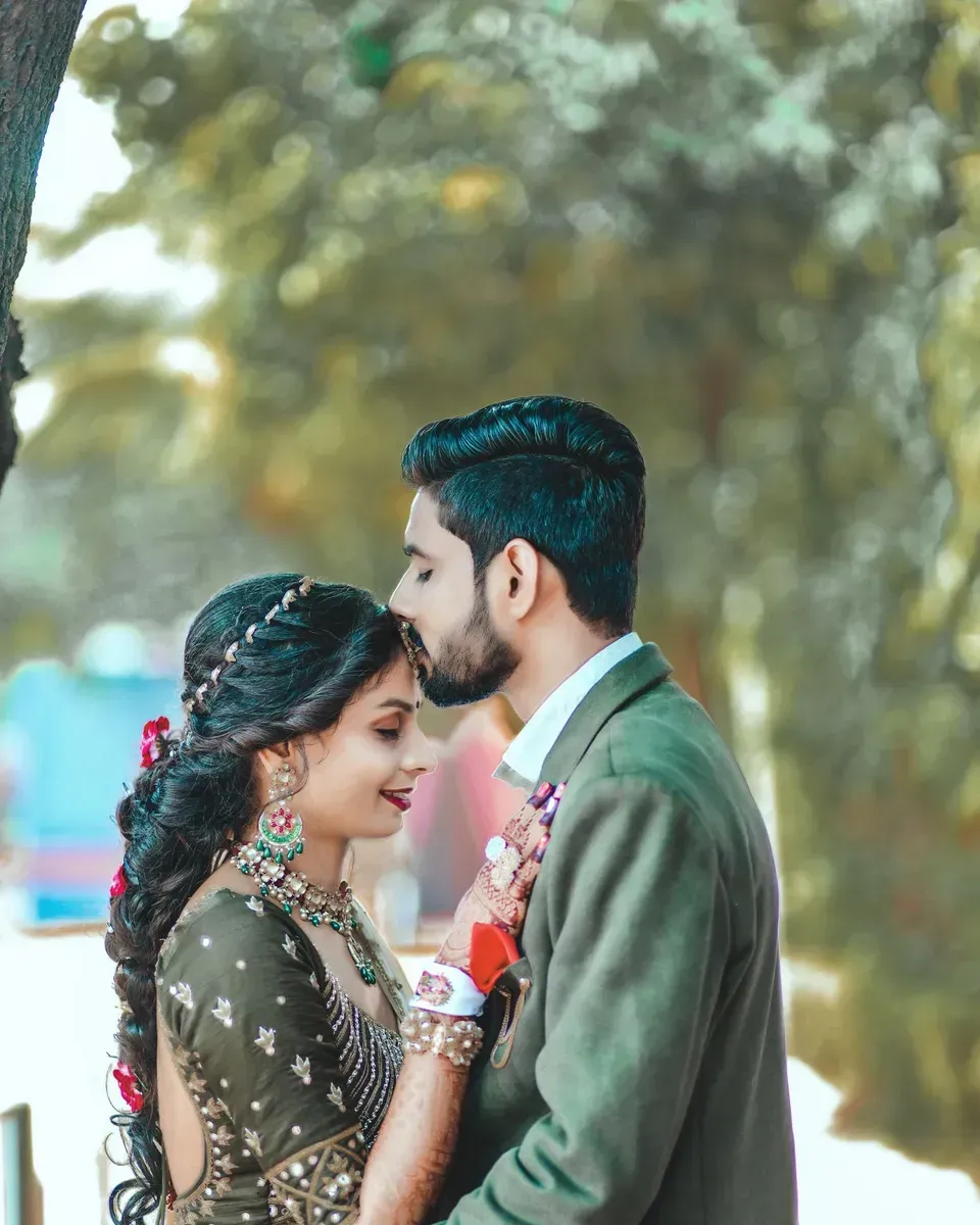 A groom kisses his bride on the forehead under green trees.